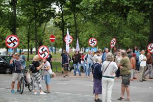 Querdenken-Protest. Viele Menschen stehen beieinander, einige halten leicht durchgestrichene Hakenkreuzschilder in die Höhe.