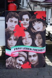 Ein Plakat mit der Aufschrift "Frau Leben Freiheit" mit den iranischen Farben. Drum herum sieht man die Gesichter einiger der ermordeten Menschen.